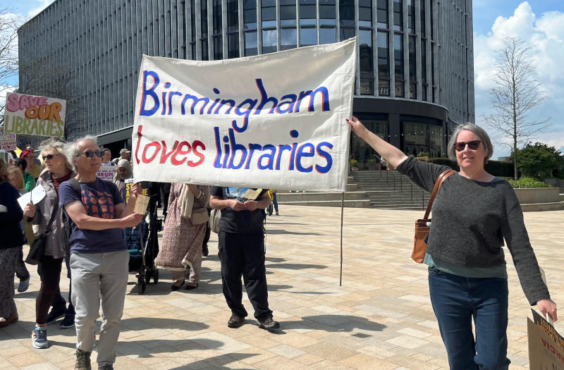 Phil holding up one end of a "Birmingham loves libraries" banner, with Nicky of Stirchley Art Room holding the other. A modern city centre building is in the background