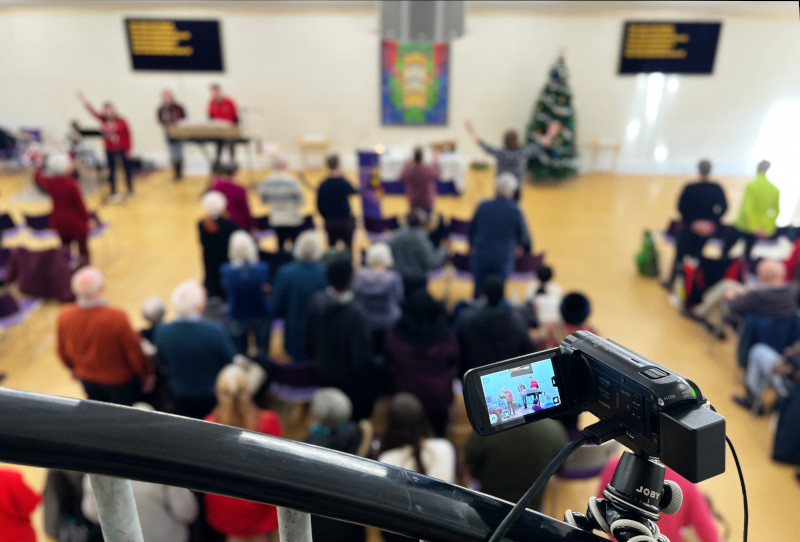 In the foregound, a camcorder mounted on a stair rail, with the focus on its display screen. In the background, out of focus, looking down on a church service taking place in the hall below