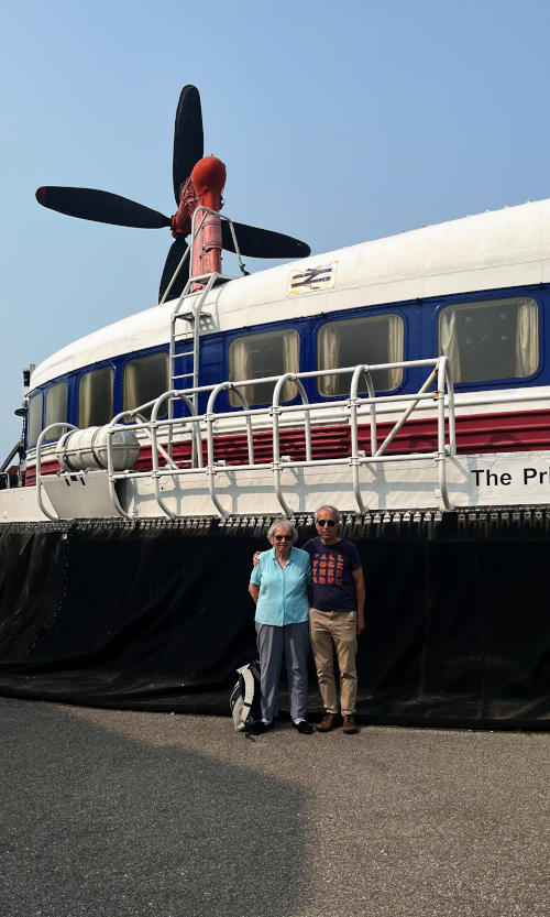 Miriam and Phil standing in front of a section of a huge hovercraft: our heads just reach to the top of its skirt. A propeller is visible above the roof of the craft