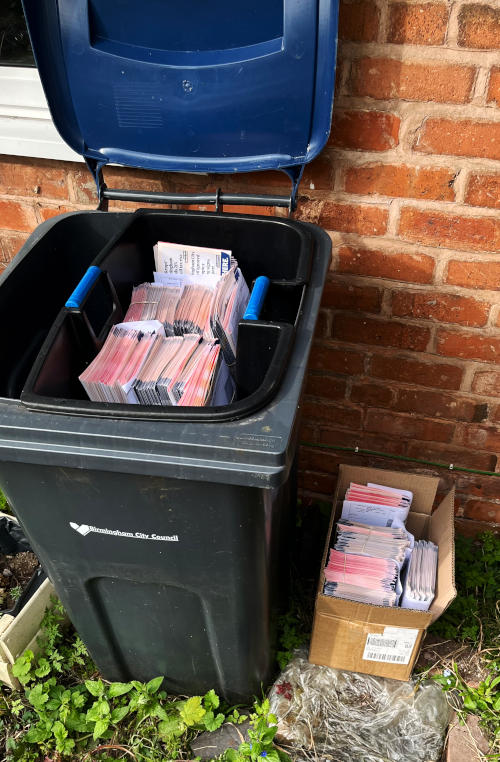 Bundles of leaflets in the paper pod at the top of an open recycling bin in front of a wall