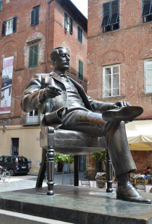 Huge statue of Giacomo Puccini seated in a chair, with a tall house behind