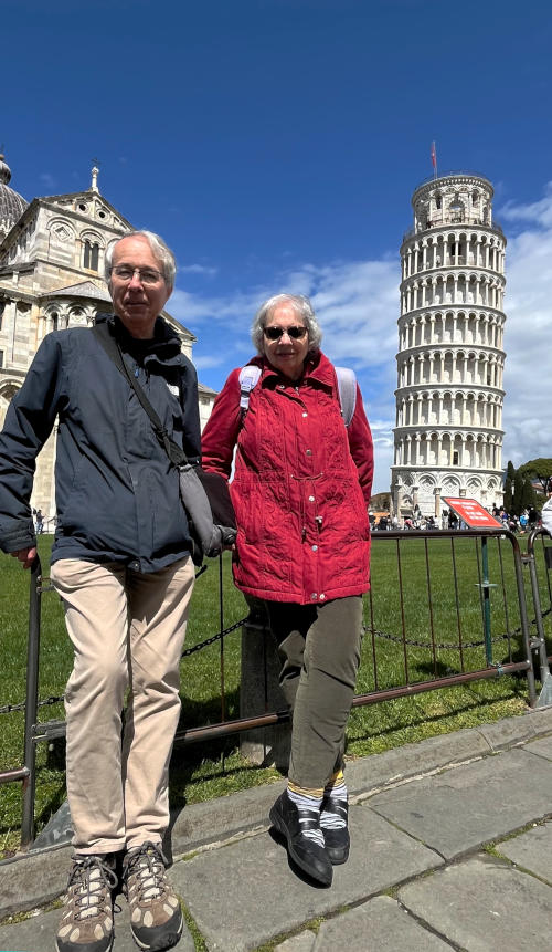 Phil and Miriam standing against a low metal fence with the Leaning Tower of Pisa in the background