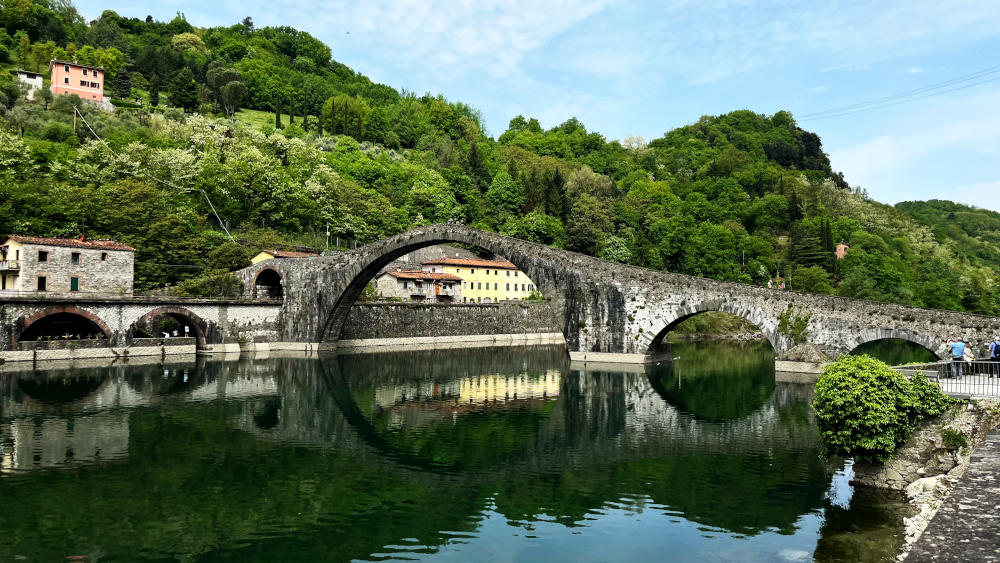 A long and slender arch bridge with a wide central span crossing a river with wooded hills on the far side