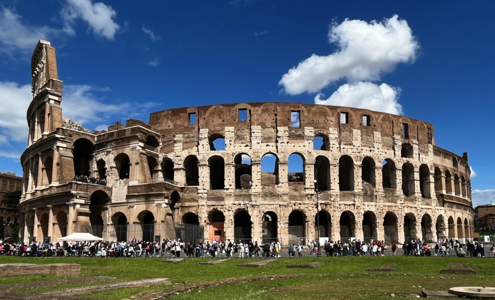 Outside view of the Colosseum in sunshine, showing its arches, with many tourists walking around in front