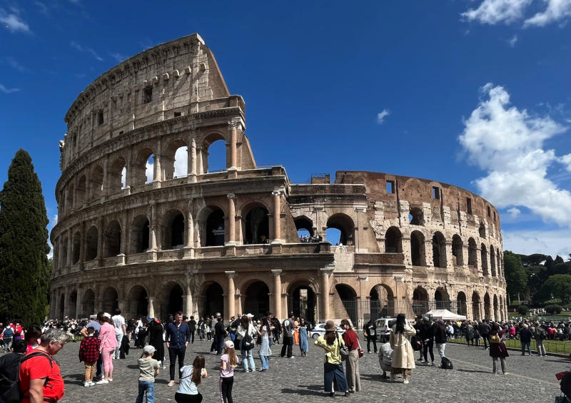 Outside view of the Colosseum in sunshine, showing its arches, with many tourists in the open space in front