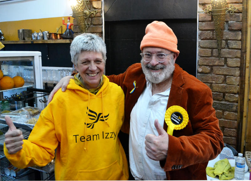 Izzy Knowles wearing a yellow Team Izzy sweatshirt standing next to campaigning journalist John Sweeney, both facing the camera with thumbs up