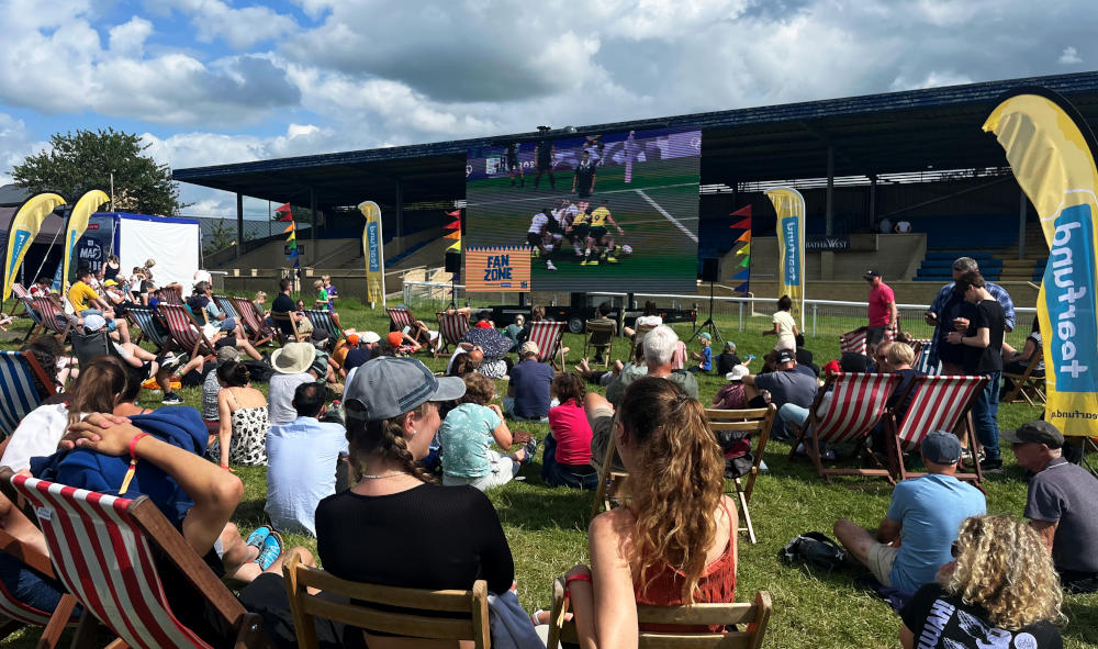 A crowd sitting in deckchairs and on the grass, in sunshine, watching rugby sevens in the Olympic Games on a giant screen