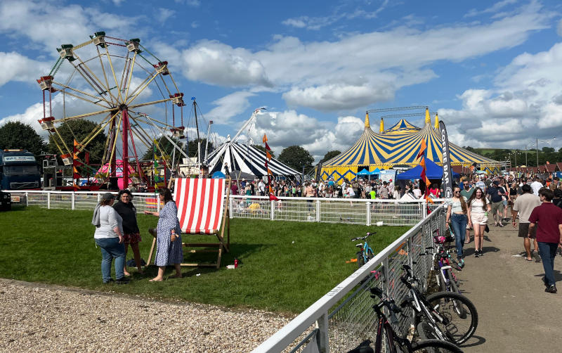 Looking towards a large field behind a low fence, within which is a Ferris wheel, two huge marquees and smaller stalls with many people, all in sunshine