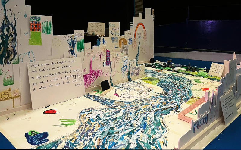 Cardboard model of a city, with artwork depicting some Christian themes, and artwork representing a river on the floor