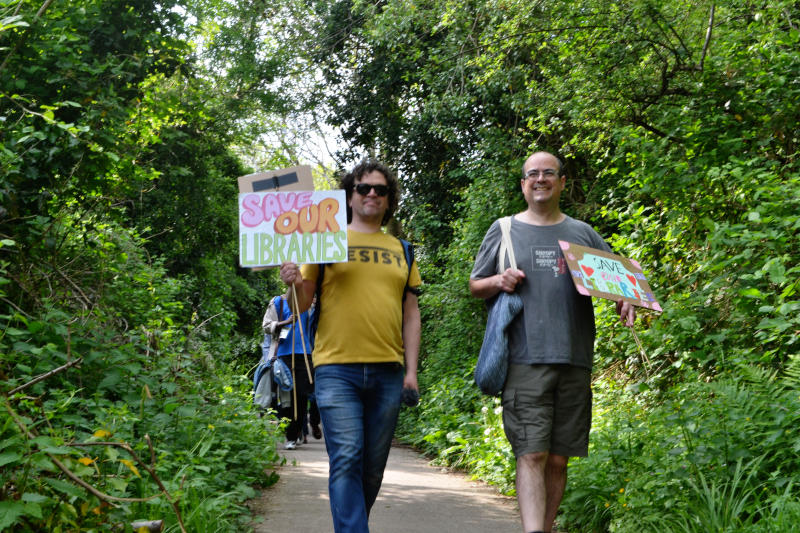 Two men walking towards the camera along a tree-lined path holding "Save Our Libraries" placards