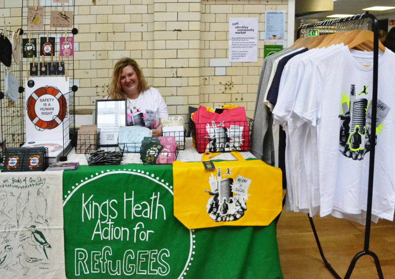 Kings Heath Action for Refugees market stall at the indoor Stirchley Community Market, with the tiled wall of Stirchley Baths behind. The stall is selling refugee themed T-shirts, bags and other products